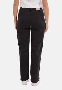 Persona che indossa pantaloni neri a gamba dritta e sneakers bianche, vista da dietro su uno sfondo bianco.