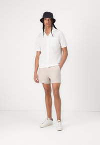 Chemise blanche à manches courtes avec un poche, shorts beiges et chapeau seau noir. Des baskets blanches complètent la tenue sur un fond uni.