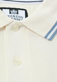 Kerman neulepolo kauluksessa kaksi nappia ja siniraitainen reunus, jossa näkyy "Weekend Offender" -labeli kauluksen sisäpuolella.