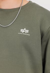 Olivovo zelená mikina so elastickým golierom a manžetami. Na prednej strane sa nachádza biele logo "Alpha Industries" a geometrický dizajn.