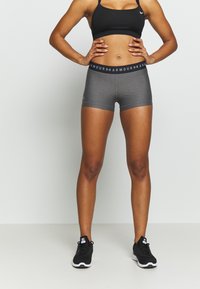 Shorts sportivi grigi con una vita nera che mostra il testo "ARMOUR." Abbinati a un reggiseno sportivo nero e scarpe da ginnastica nere.