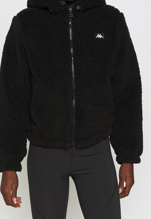 Sort lynlåsfleecejakke med hætte, båret over sorte leggings, viser tekstureret stof og lille hvidt logo på brystet.