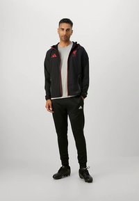 Zwarte sportjas met rode accenten en Liverpool-logo, gecombineerd met een grijs T-shirt en zwarte broek, afgemaakt met zwarte voetbalschoenen.