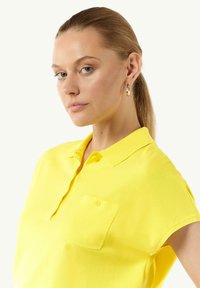 Gelbes Poloshirt aus weichem Stoff, mit einem Kragen, kurzen Ärmeln und einer Brusttasche mit einem einzelnen Knopfdetail.