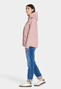 Lichtroze waterdichte jas met capuchon, voorzien van drukknoopsluitingen, twee voorzakken en een ontspannen pasvorm, gecombineerd met blauwe denim jeans en witte sneakers.