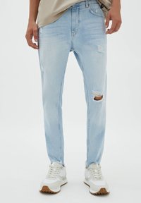 Jeans de denim azul claro con agujeros y bajos deshilachados. Diseño de corte recto con una textura desgastada. Combinados con zapatillas blancas con detalles en tono marrón.