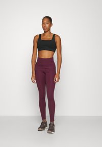 Black sports bra with square neckline, paired with high-waisted maroon leggings. Model wears gray athletic shoes. Smooth fabric, fitted design.