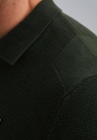 Dunkelgrünes Strick-Polohemd mit strukturierter Oberfläche, klassischem Kragen und sichtbaren Schulternähten. Der Stoff hat eine weiche, grobe Textur.