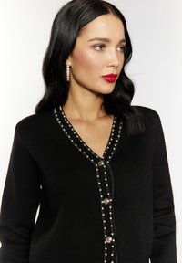 Schwarze Strickjacke mit V-Ausschnitt, verziert mit Perlen- und Metallic-Perlenakzenten entlang der Vorderseite und fünf Knöpfen; glatte Stricktextur.
