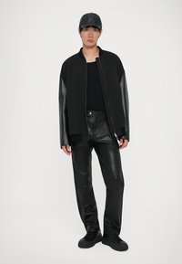 Giubbotto bomber nero con maniche in pelle, abbinato a pantaloni in pelle nera. Un berretto nero e calzature completano il look monocromatico.
