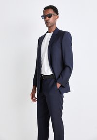 Completo blu con giacca a un bottone e pantaloni su misura, abbinato a una camicia bianca a girocollo e cintura nera. Gli accessori includono occhiali da sole.