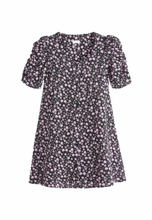 Kind steht vor einer roten Tür, trägt ein schwarzes Kleid mit pinken Blumen, gelbe Socken und schwarze Knöchelstiefel.