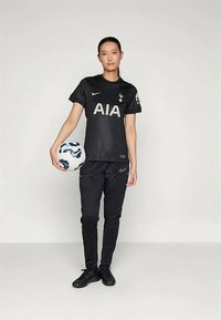 Tenue de football noire à manches courtes, arborant un logo "AIA" blanc bien visible et l'emblème de Tottenham. Assortie à un pantalon noir et des chaussures noires.