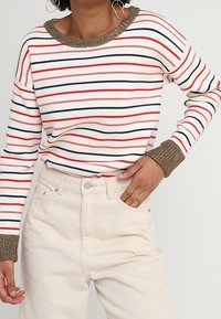 Pull à manches longues rayé en blanc, rouge et marine, avec un col et des poignets texturés marron. Associé à un jean taille haute de couleur crème.
