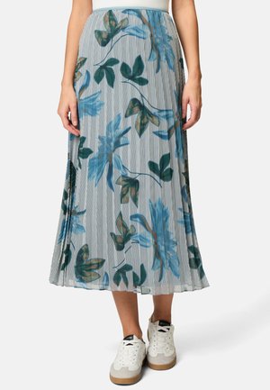 Plissé bloemenrok met een lichtblauw en grijs verticaal gestreept patroon, versierd met groene en bruine bladeren. Draagt wit sneakers.