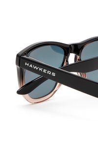 Okulary przeciwsłoneczne Hawkers z czarną i przezroczystą oprawą, ciemnymi soczewkami oraz białym logo na zausznikach. Smukły, kanciasty design z błyszczącym wykończeniem.