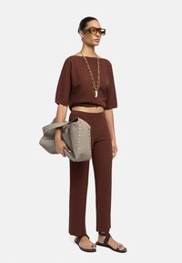Kobieta ubrana w brązowy prążkowany crop top i dopasowane spodnie, z dużym złotym naszyjnikiem, dużymi brązowymi okularami przeciwsłonecznymi, trzymająca szaro-studniowaną torebkę.