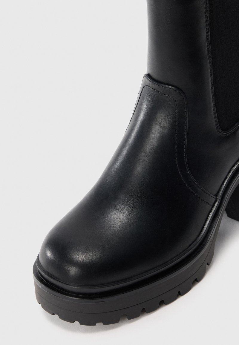 Botín de cuero negro hasta el tobillo con suela gruesa y panel lateral elástico sobre un fondo claro.