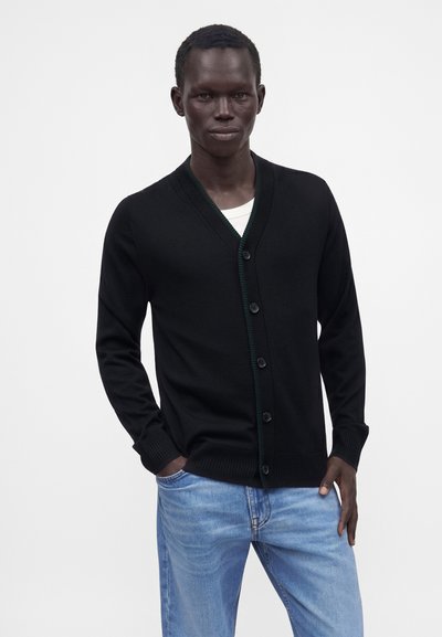 Homme portant un cardigan noir boutonné par-dessus une chemise blanche et un jean bleu clair, debout avec une main dans la poche devant un fond uni.
