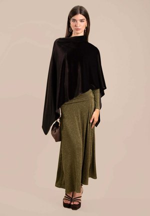 Poncho en velours noir sur une robe verte olive scintillante, associé à un sac à main marron et des talons plats foncés. Textures lisses, design fluide.