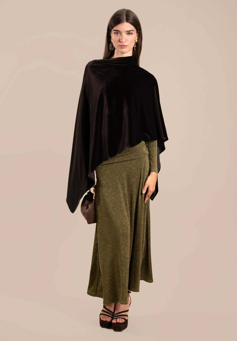 Poncho de terciopelo negro sobre un vestido verde oliva brillante, combinado con un bolso marrón y tacones de plataforma oscuros. Texturas suaves, diseño fluido.