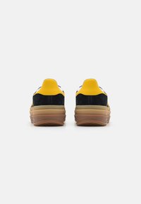 Zapatillas con una parte superior de ante amarilla y azul marino, una suela de goma texturizada y una entresuela en capas. La vista trasera muestra un talón redondeado y un contraste de colores.