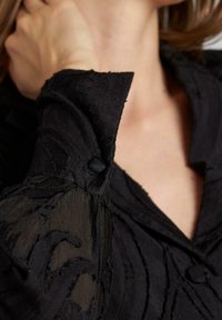Zwarte stoffen kleding met textuur en geborduurde patronen; heeft een kraag, knoopdetails en doorschijnende mouwsecties.