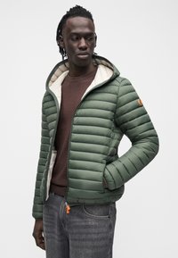 Vihreä toppatakki, jossa vaakasuora tikattu kuvio, beige vuori, säädettävä huppu ja oranssi vetoketjun vetimen. Päällä ruskea toppi.