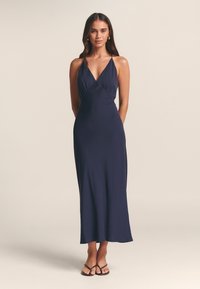 Tamsiai mėlyna, be rankovių maxi suknelė su gilia V formos iškirpte ir surinkta detalė. Lygus audinys ir laisvai krentanti medžiaga, derinama su juodomis šlepetėmis.