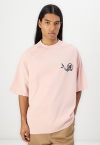 Lyse rosa oversized t-skjorte laget av bomull, med rund hals og korte raglan ermer. Viser et fargerikt brodert design på brystet.