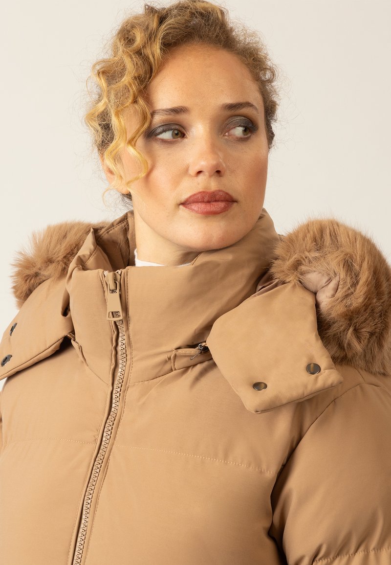 Beige Pufferjacke mit einem mit Fell gefütterten Kapuze, ausgestattet mit einem durchgehenden Reißverschluss, Druckknopf-Verzierungen und einer glatten, strukturierten Oberfläche.