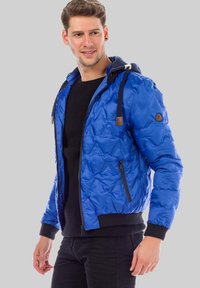 Blue padded jacket with a hood, quilted texture, black accents, front zipper, and elastic cuffs. Worn over a black knit sweater.