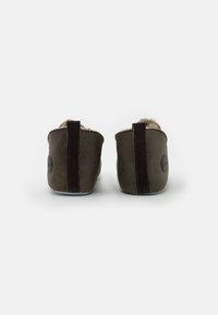 Brune ruskindslippers med en blød pelsfor, har et lodret mørkebrunt accent bagpå og et cirkulært præget logo på siden.