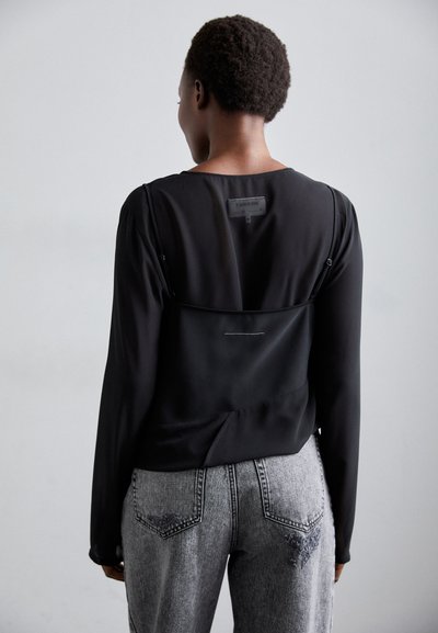 Blusa nera a maniche lunghe con una texture liscia, caratterizzata da un design a strati. Sul retro presenta dettagli di cucitura e un'etichetta. Indossata con jeans in denim grigi.