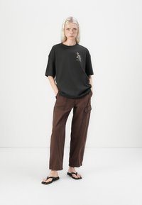 Black oversized t-shirt with a graphic design on the chest, paired with loose brown cargo pants and black flip-flops.