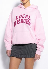 Light pink hoodie with "LOCAL HEROES" in bold burgundy letters across the chest. Features a front pocket and a loose, comfortable fit.