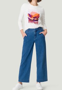 Chemise blanche à manches longues avec l'inscription « MINDSET IS EVERYTHING », associée à un jean large en denim bleu et des baskets blanches. Design simple.