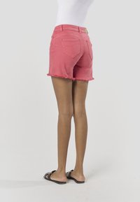 Różowe szorty z denimu z postrzępionymi brzegami i tylnymi kieszeniami. Talia ozdobiona jest markową metką. Noszone z czarnymi klapkami.