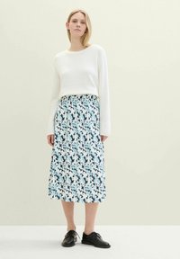 Eine Frau in einem weißen Langarmpullover, einem blau-weißen, blumigen, gefaltenen Midikleid und schwarzen Schnürschuhen steht vor einem einfarbigen Hintergrund.