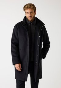 Cappotto lungo in misto lana nero con colletto alto, chiusura a bottoni e cerniera, e due tasche frontali. Fodera interna imbottita, che offre calore.