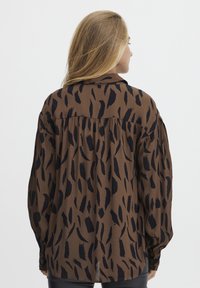 Brun skjorte med knapper og et sort abstrakt mønster, lange ærmer, plisseret rygdetalje og løs pasform. Glat stofstruktur.
