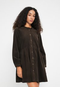 Donna con capelli ricci che indossa un abito camicia lungo in velluto a coste marrone, in piedi su uno sfondo bianco uniforme.