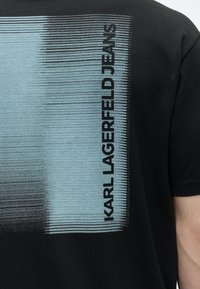 Μαύρο T-shirt με κάθετες μπλε γραμμές με βαθμιαία απόχρωση και το λογότυπο "KARL LAGERFELD JEANS" εκτυπωμένο κάθετα στην πλάτη κοντά στον ώμο.