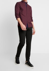 Man in een bordeauxrood overhemd met knopen, zwarte slim-fit broek en zwarte sneakers met witte zolen, staand tegen een effen witte achtergrond.