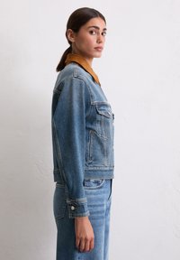 Veste en denim bleu clair avec un col en suède marron. Elle comprend deux poches poitrine, des fermetures à boutons et une silhouette ajustée.