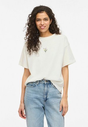 Femme aux cheveux bouclés portant un T-shirt blanc oversize avec une petite broderie florale, rentré dans un jean taille haute bleu clair.