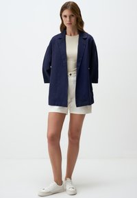 Blazer in lino blu navy con vestibilità rilassata, rever a punta e maniche lunghe, abbinato a pantaloni corti bianchi a vita alta e scarpe da ginnastica bianche.