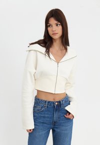 Gekürzte weiße Zip-Hoodie mit weitem Kragen, hergestellt aus weichem Stoff. Kombiniert mit hochgeschnittenen blauen Jeans.