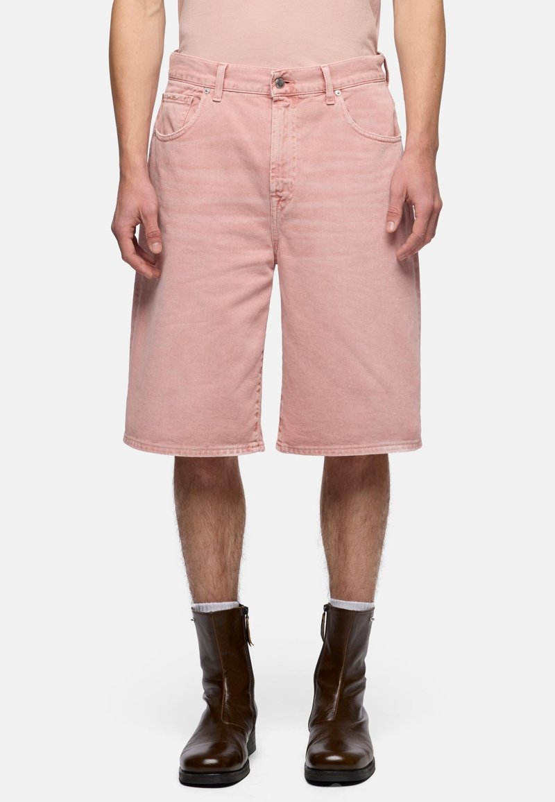 Uomo indossa pantaloncini di jeans rosa sbiadito fino al ginocchio, calzini chiari e stivali alla caviglia in pelle marrone scuro, in piedi contro uno sfondo bianco.