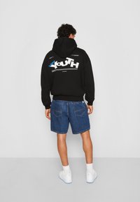 Hoodie nero con grafica "Youth" bianca e blu sulla schiena, abbinato a pantaloni corti di jeans blu. Il modello indossa sneakers bianche e calzini.